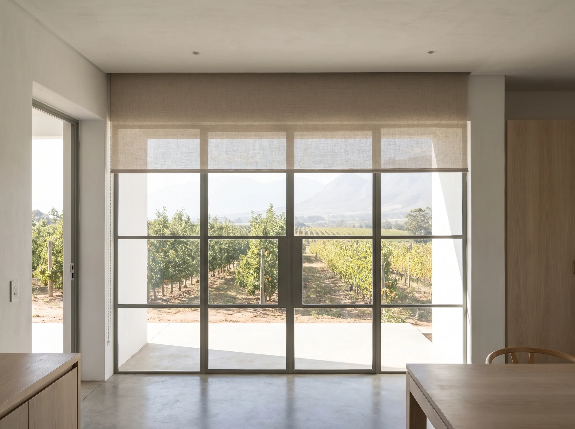 Roller blind dropping from a concealed ceiling slot over steel-framed glazing in a new Simondium farm cottage, orchard view beyond